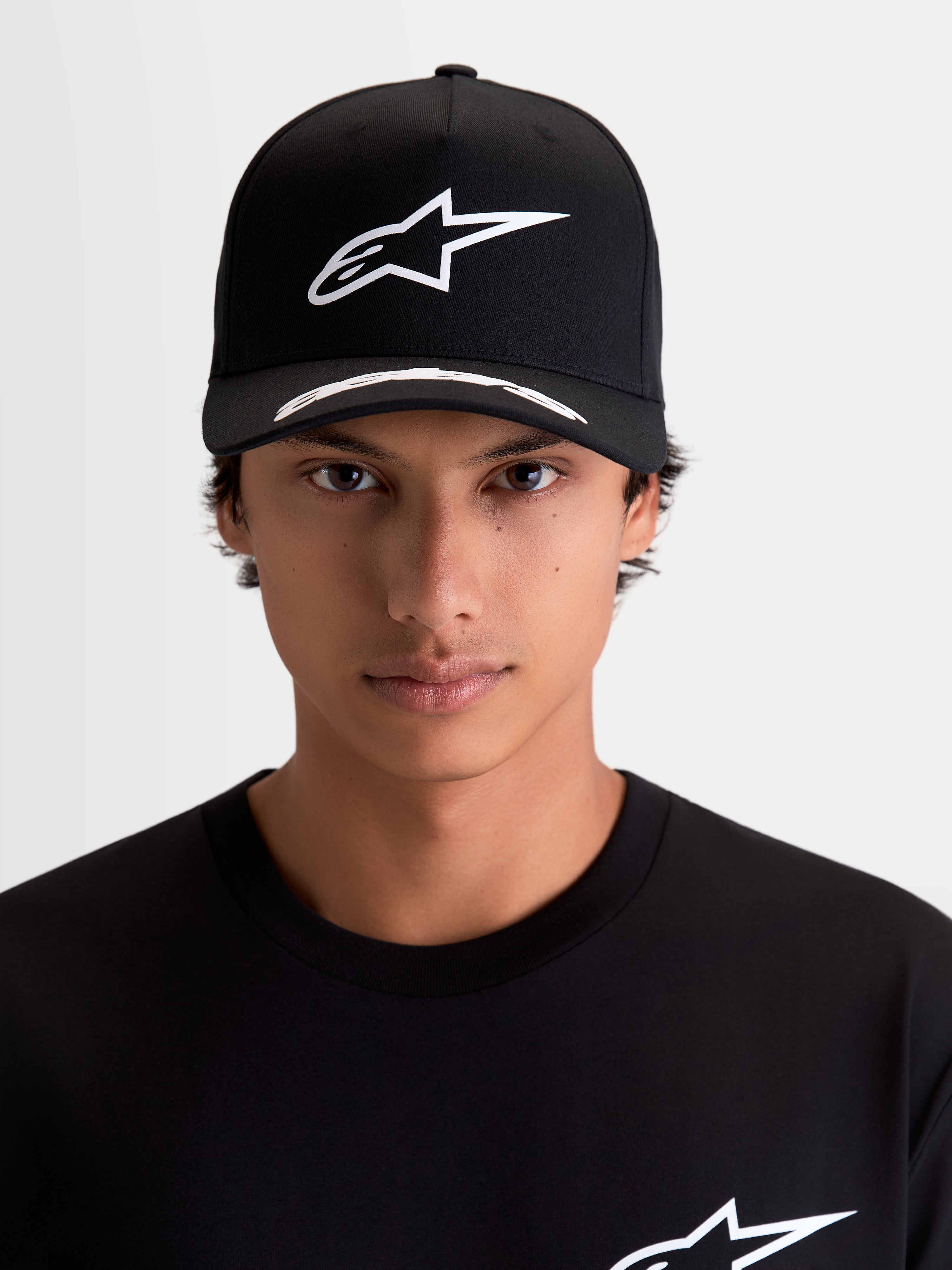 Alpinestars Arclight, gorra de estilo motorsport y sportswear, negra y blanca, vista frontal en modelo, con logotipo Astars en relieve blanco sobre corona estructurada, visera curva con detalles y tejido de sarga premium.