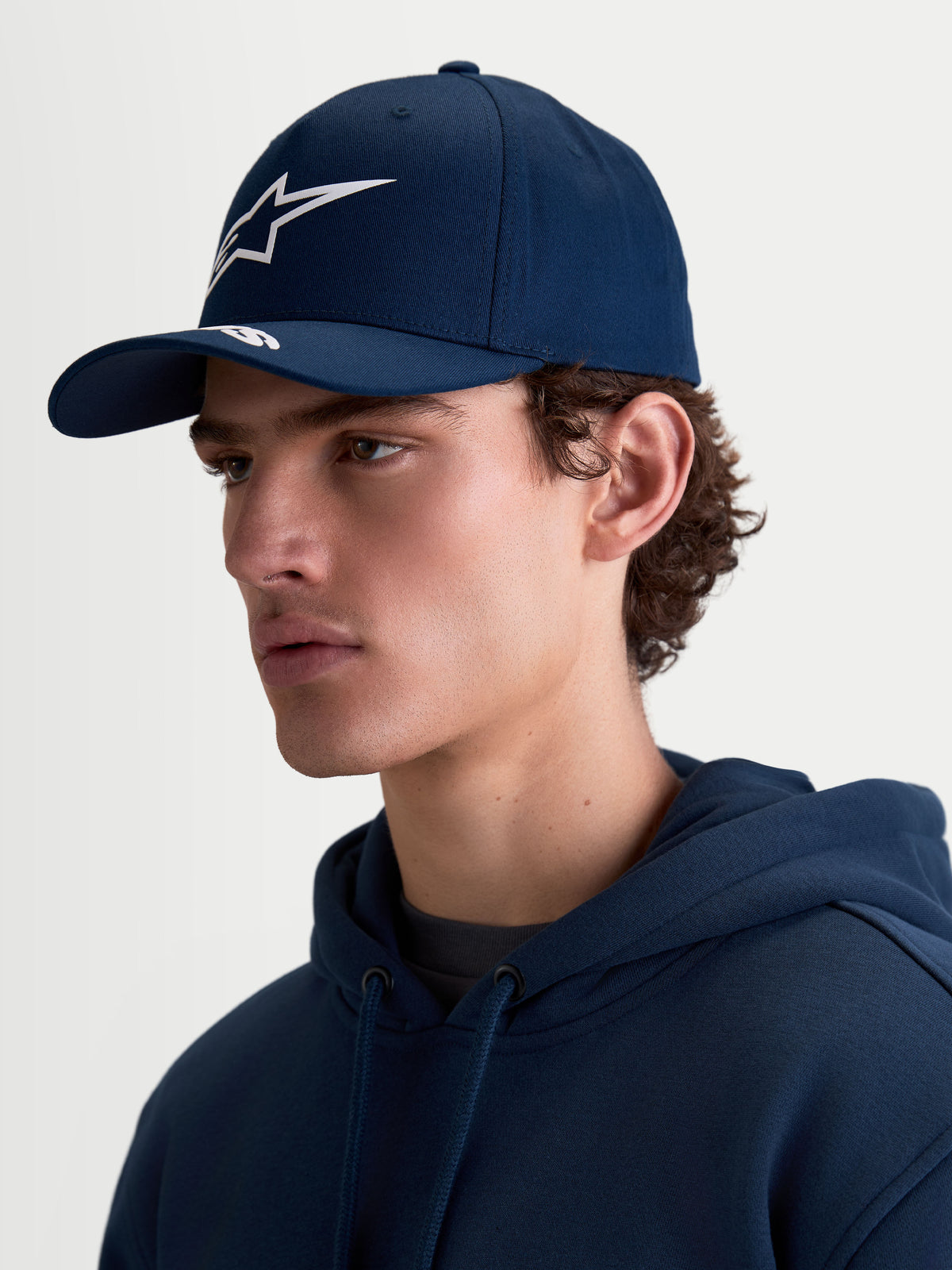 Alpinestars Arclight, gorra de béisbol estilo sportswear para hombre y mujer, azul marino y blanco, logotipo Astars en relieve, visera curva, corona estructurada, tejido de sarga de algodón, vista de perfil lateral en modelo.