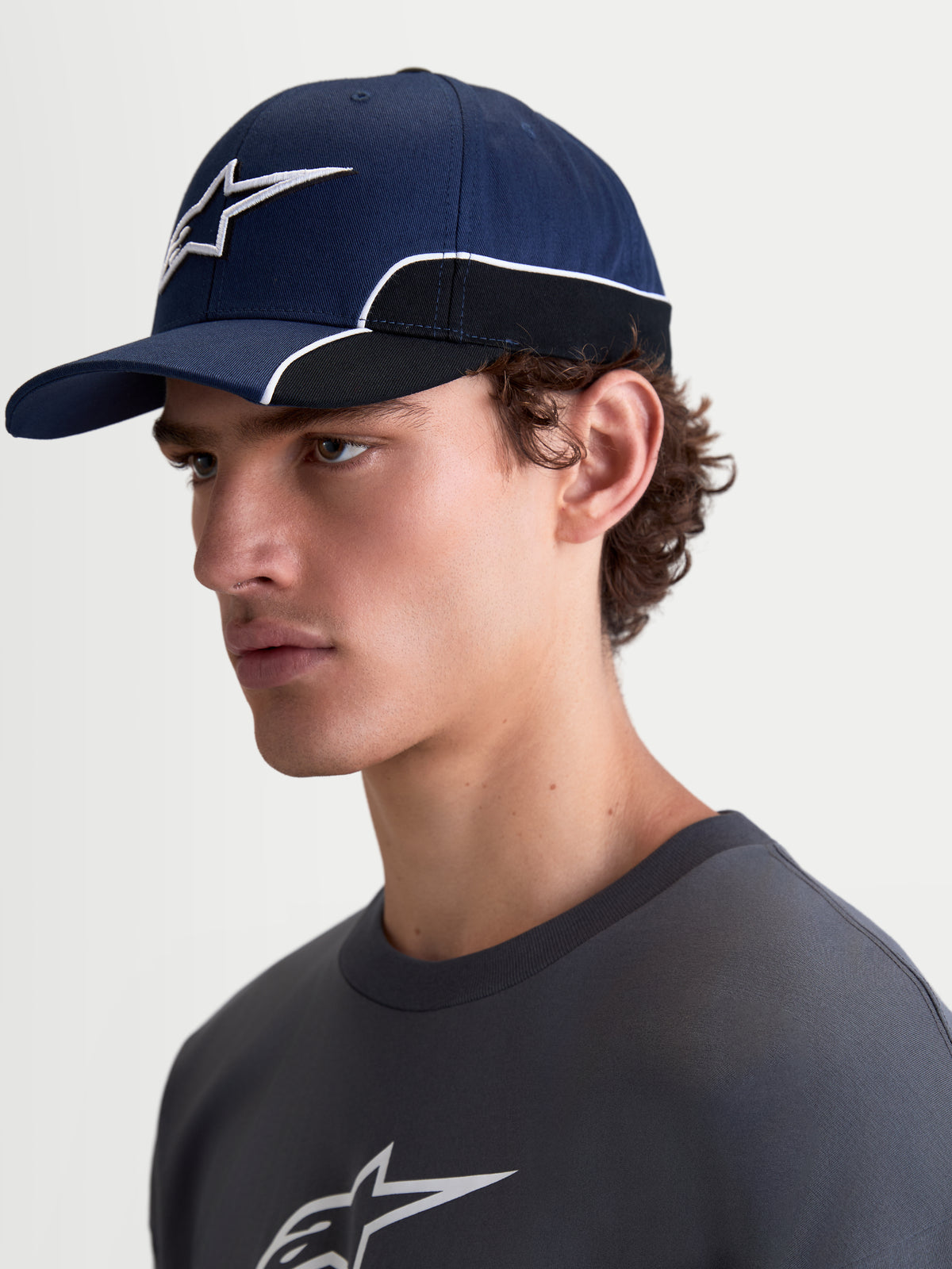 Alpinestars gorra 500 Snapback, gorra de béisbol estilo sportswear, azul marino y negra, confeccionada en algodón resistente, cuenta con visera curva, logotipo Astars bordado en 3D y ribetes blancos en contraste, vista lateral en modelo.