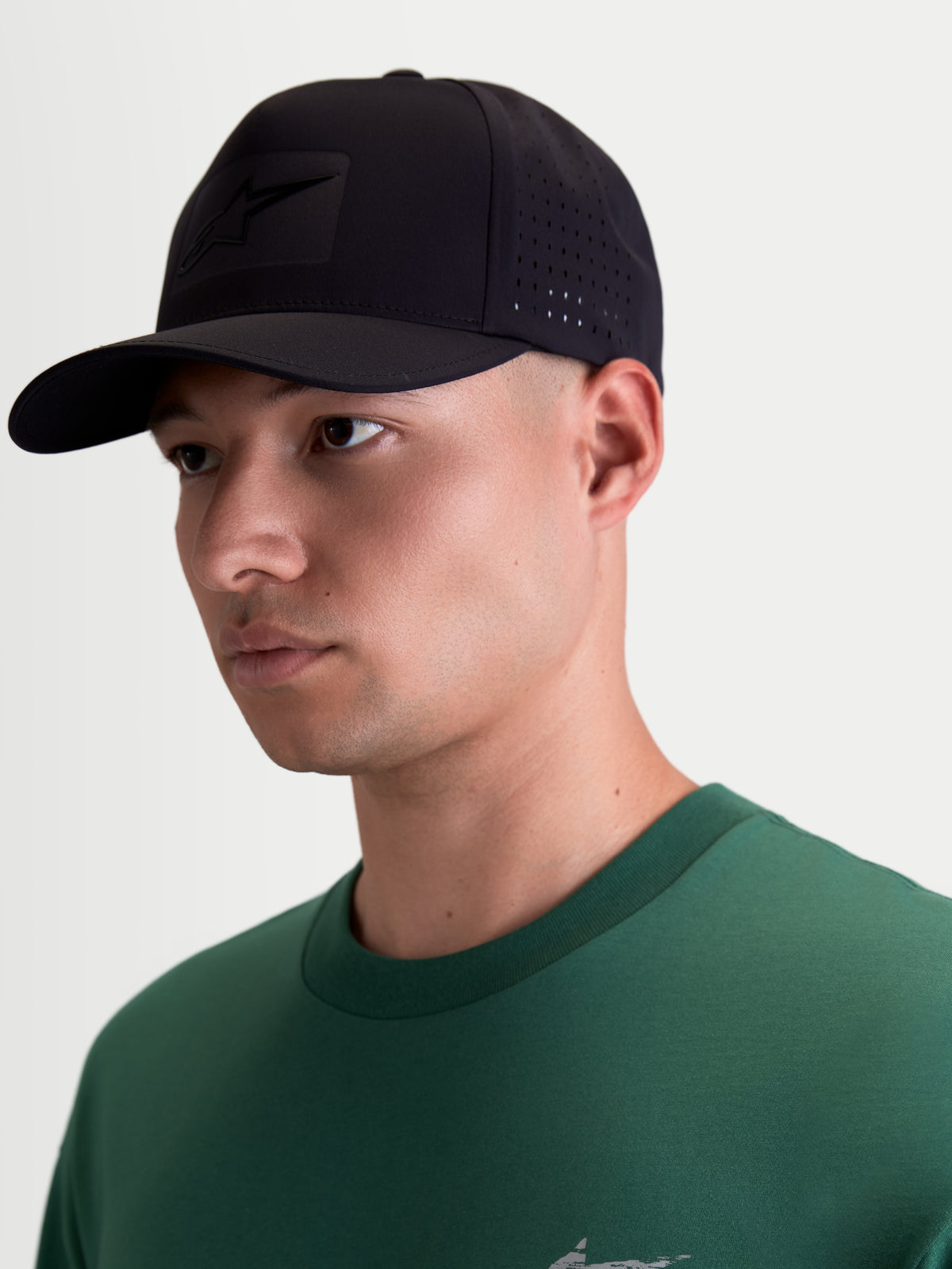 Alpinestars Algo Perf Ajustable, gorra deportiva de estilo motorsport, vista en modelo, color negro, visera curva, tejido técnico con perforaciones laterales para ventilación y logotipo Astars tonal con soldadura sónica.