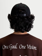 Alpinestars Gorra Retro Snapback, gorra snapback de estilo ropa deportiva, negra y verde, vista trasera en modelo con cierre ajustable y detalles de ojales verdes, combinada con una camiseta marrón con el manifiesto de marca One Goal. One Vision.