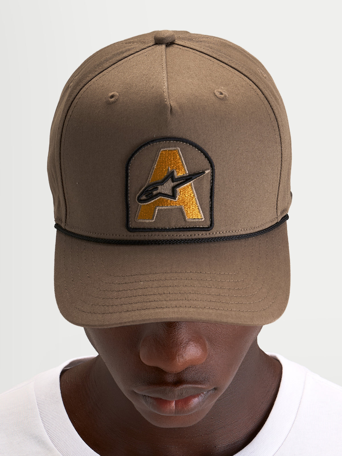 Gorra Alpinestars Acme Snapback, gorra de sportswear con visera curva, marrón nuez con parche bordado del logotipo Astars en el frontal y ribete de cuerda negro, vista frontal en modelo
