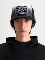 Alpinestars Gorra Gravel, gorra de estilo trucker, negra y blanca, vista frontal en modelo con visera curva, malla trasera transpirable y parche tejido Racing Ahead con detalles de textura de grava.
