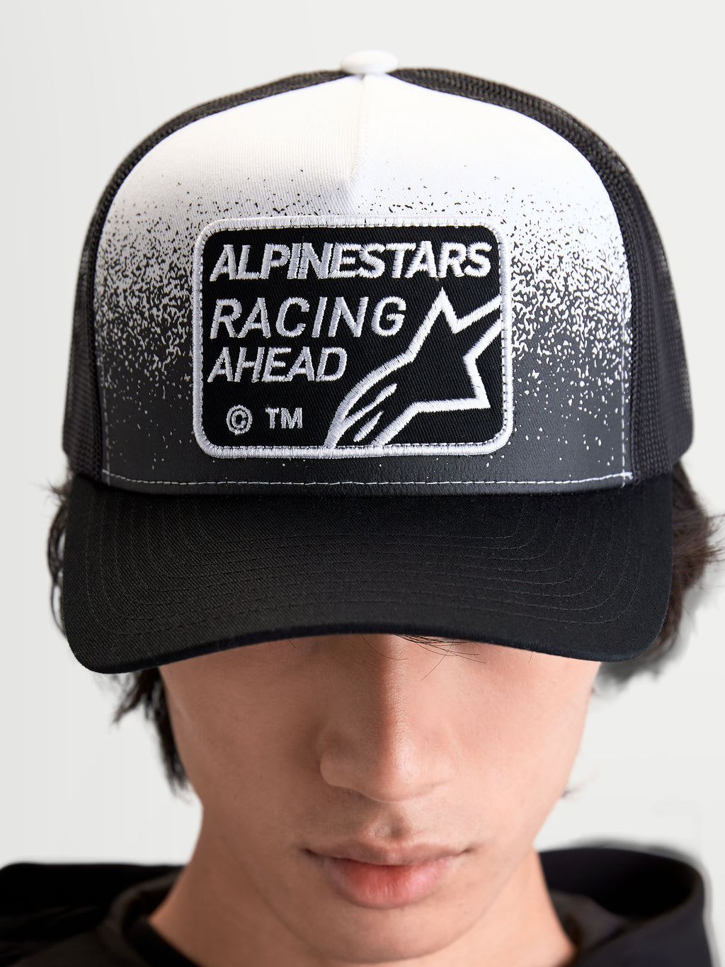 Alpinestars Gorra Gravel, gorra de estilo trucker de estilo sportswear, negra y blanca, con parche frontal tejido Alpinestars Racing Ahead y textura de grava, corona estructurada y visera curva, vista frontal en modelo.