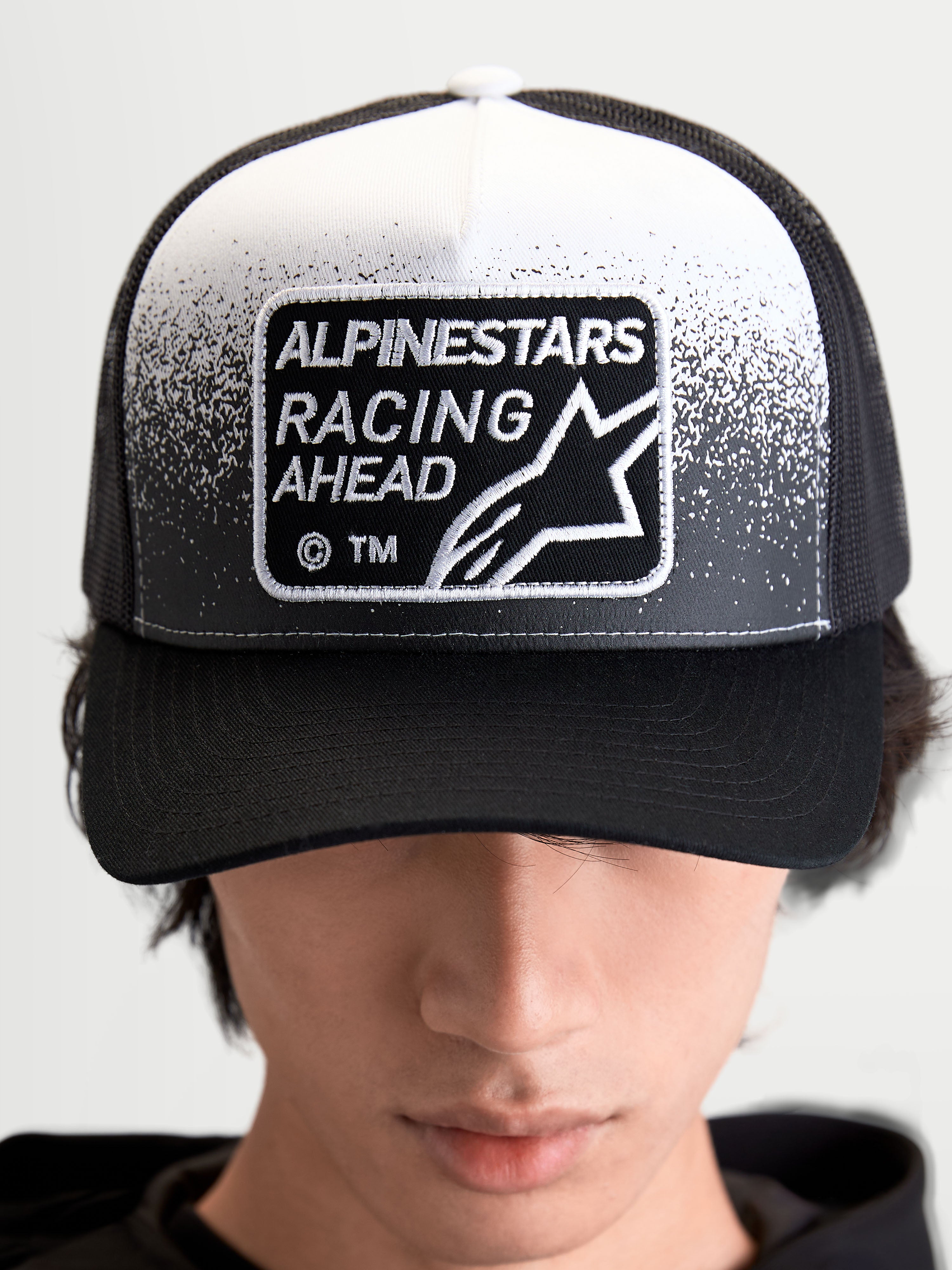 Alpinestars Gorra Gravel, gorra de estilo trucker de estilo sportswear, negra y blanca, con parche frontal tejido Alpinestars Racing Ahead y textura de grava, corona estructurada y visera curva, vista frontal en modelo.