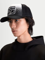 Alpinestars Gorra Gravel, gorra de estilo trucker mostrada en modelo, negra y blanca, con corona estructurada, visera curva, malla trasera transpirable y parche tejido frontal con gráficos de textura de grava y branding Alpinestars Racing Ahead.