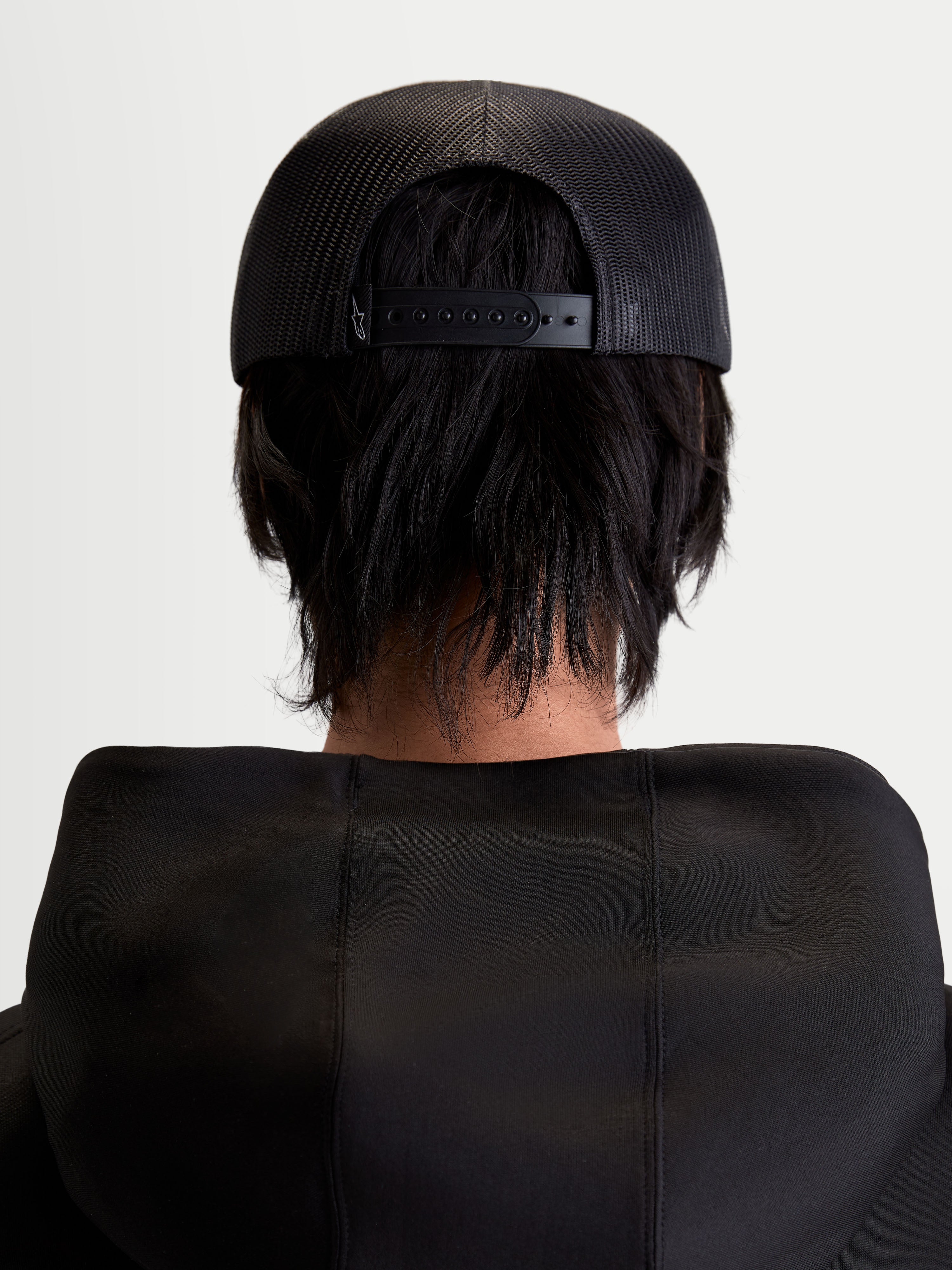 Alpinestars Gorra Gravel, gorra de estilo trucker unisex, color negro, vista trasera en modelo mostrando la malla de poliéster transpirable y el cierre ajustable a presión.