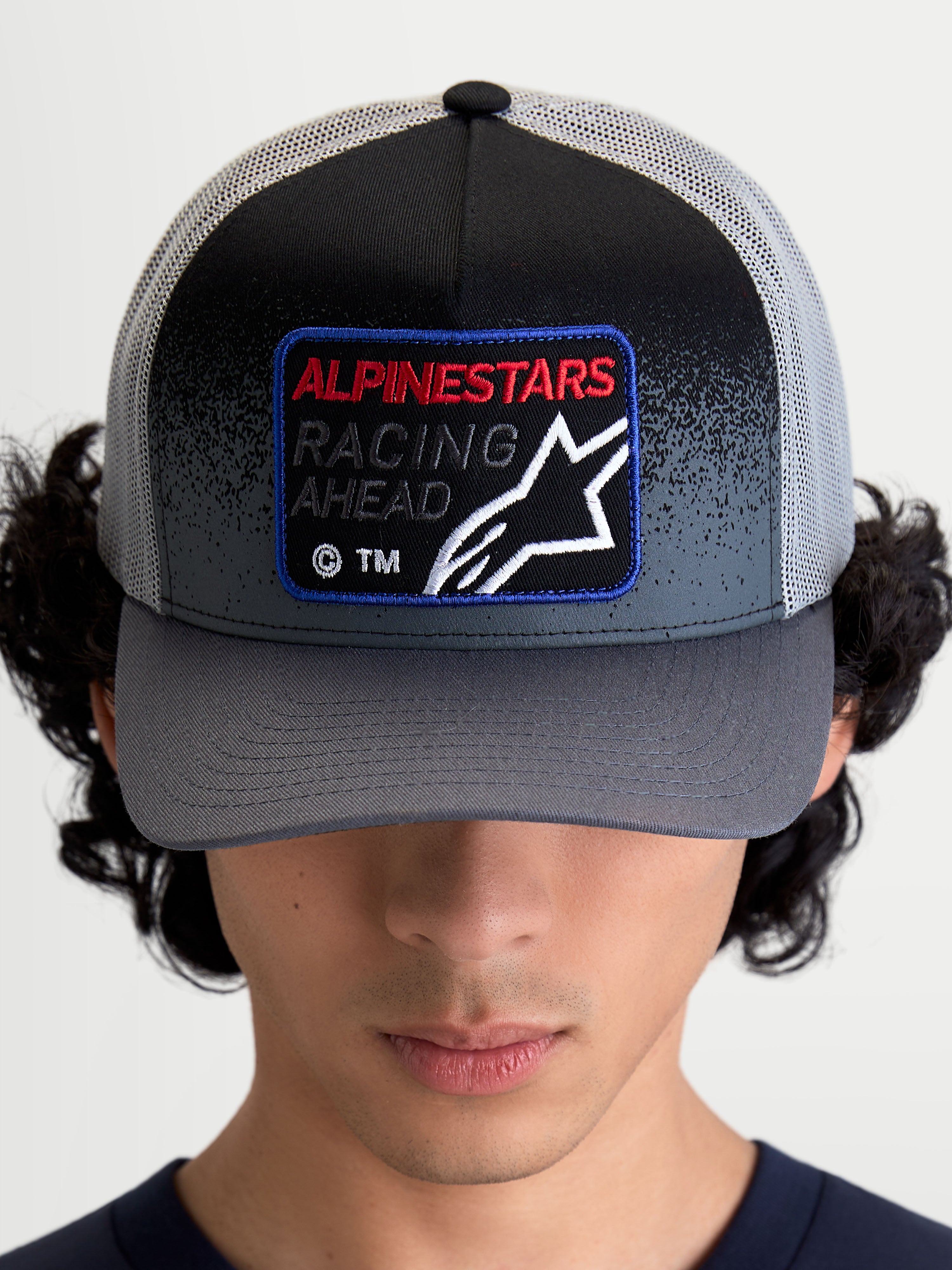 Alpinestars Gorra Gravel, gorra de estilo trucker para ropa deportiva, gris oscuro y negra, visera curva, parche frontal tejido con texto Racing Ahead y logotipo Astars, vista frontal en modelo