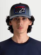 Alpinestars Gorra Gravel, gorra de malla para hombre y mujer, gris oscuro y negra, mostrada en modelo. Incluye corona estructurada, visera curva y parche frontal tejido con diseño Alpinestars Racing Ahead y detalles con textura de grava.