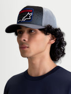 Gorra Gravel Alpinestars, gorra de estilo casual puesta en modelo, gris oscuro y negro con malla trasera gris claro, corona estructurada, visera curva y parche frontal tejido con texto Alpinestars en rojo, ideal para un estilo cotidiano inspirado en el automovilismo