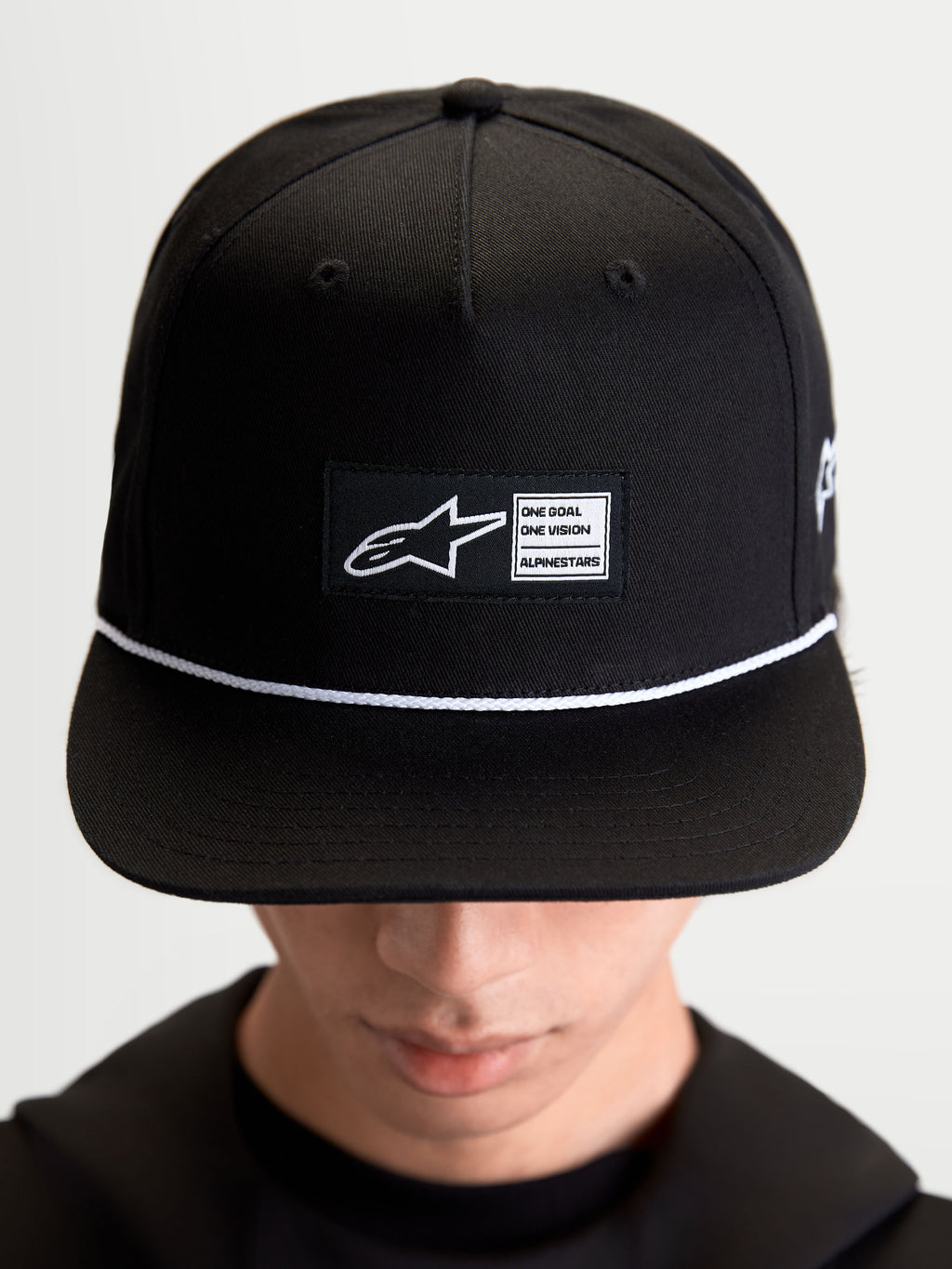 Alpinestars Gorra Label Snapback, gorra de visera plana estilo sportswear, unisex, negra y blanca, corona estructurada de sarga de algodón con parche tejido frontal y cordón blanco de contraste, mostrada en modelo.