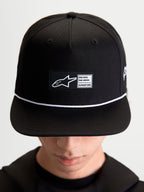 Alpinestars Gorra Label Snapback, gorra de visera plana estilo sportswear, unisex, negra y blanca, corona estructurada de sarga de algodón con parche tejido frontal y cordón blanco de contraste, mostrada en modelo.
