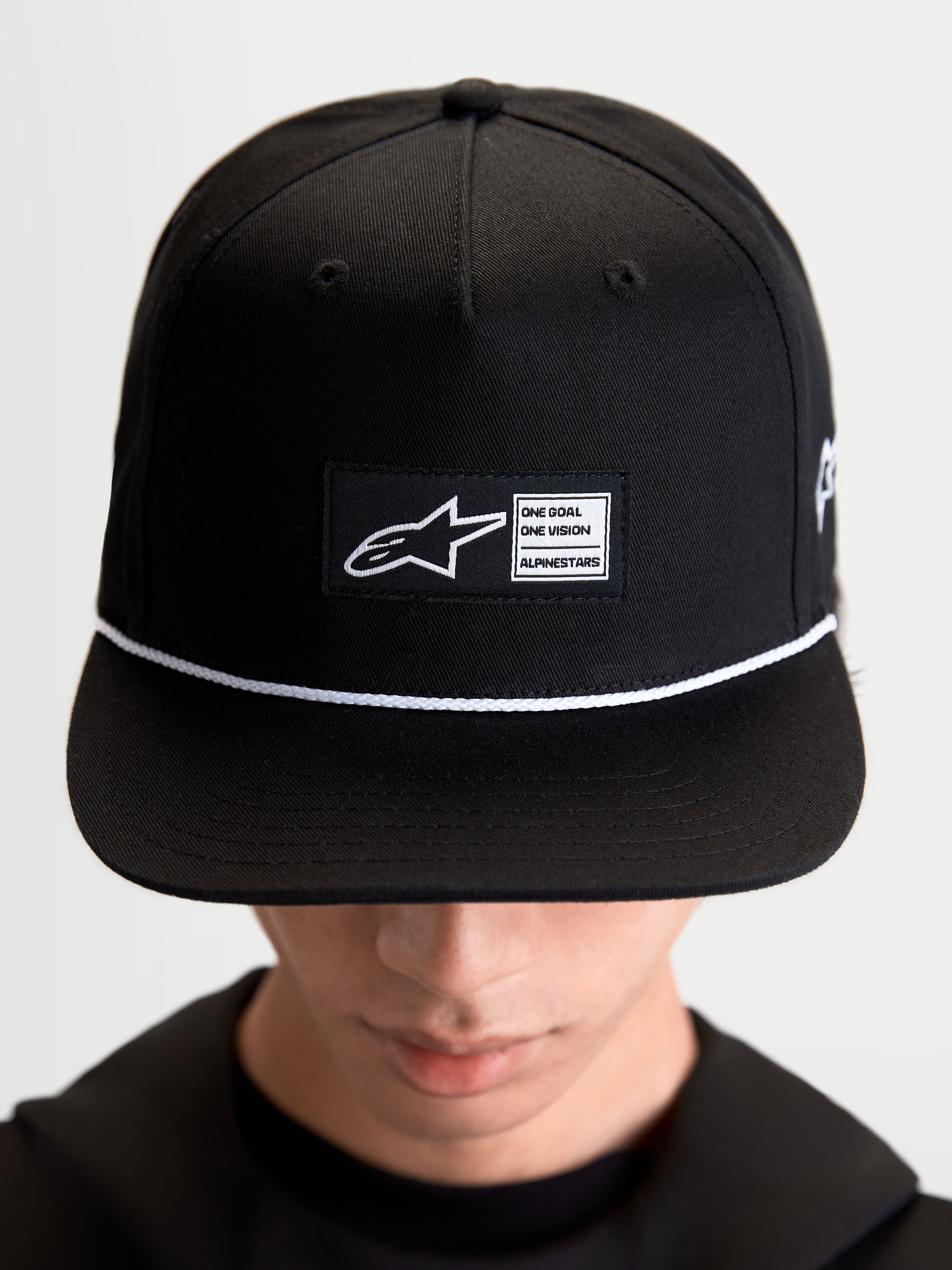 Alpinestars Gorra Label Snapback, gorra de visera plana estilo sportswear, unisex, negra y blanca, corona estructurada de sarga de algodón con parche tejido frontal y cordón blanco de contraste, mostrada en modelo.