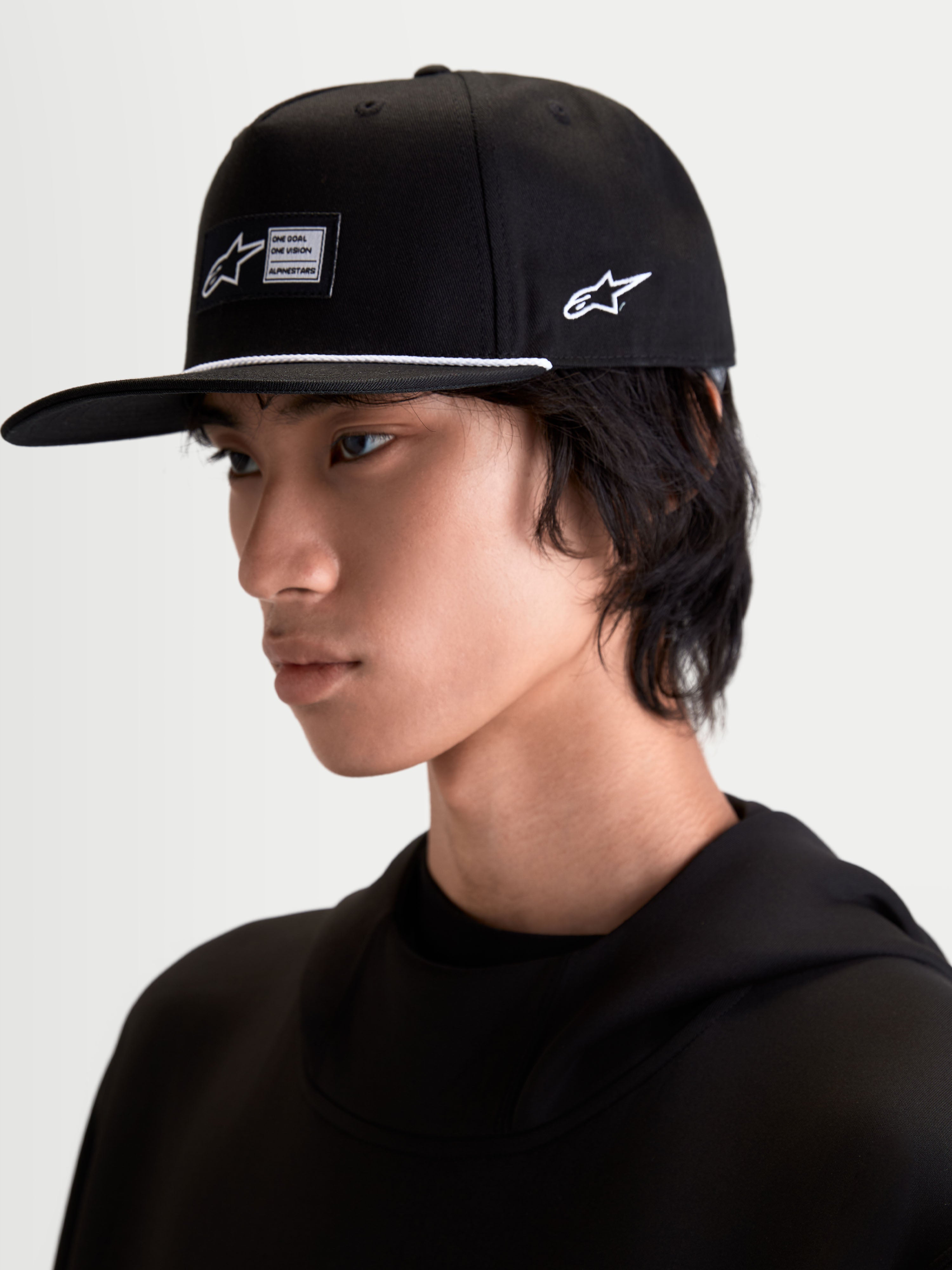 Alpinestars Gorra Label Snapback, gorra de visera plana y corona estructurada, color negro con cordón de acento blanco y etiqueta frontal tejida, mostrada en modelo para un look sportswear inspirado en el automovilismo