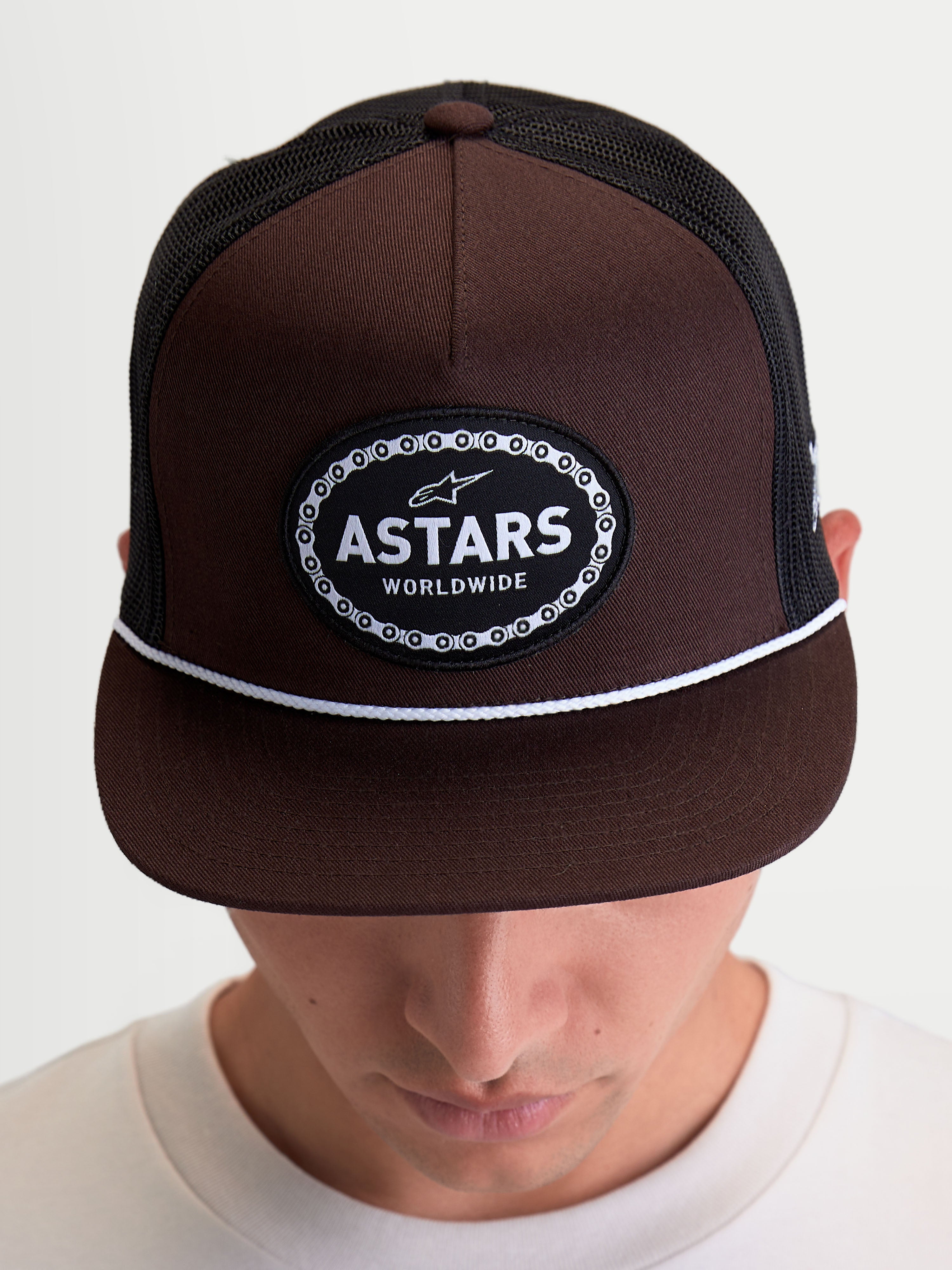 Alpinestars Hotlink Trucker Hat, Motorsport-inspired Sportswear Cap shown on model, Brown and Black, featuring a structured crown, flat bill with a white cord detail, and a front Astars Worldwide logo patch with a bike chain border.