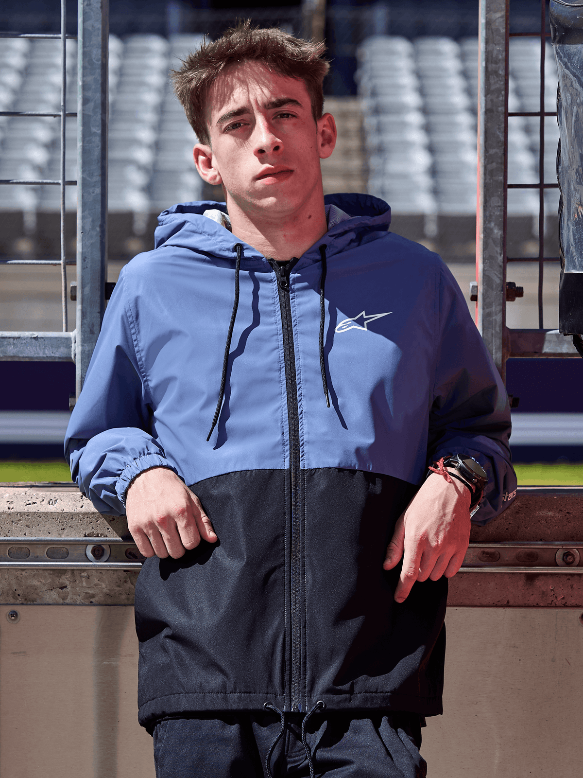 Chaqueta cortavientos Alpinestars Speeder, azul pizarra y negro, cortavientos con capucha y diseño de bloques, logo blanco en el pecho, prenda de estilo de vida casual usada en un circuito de carreras
