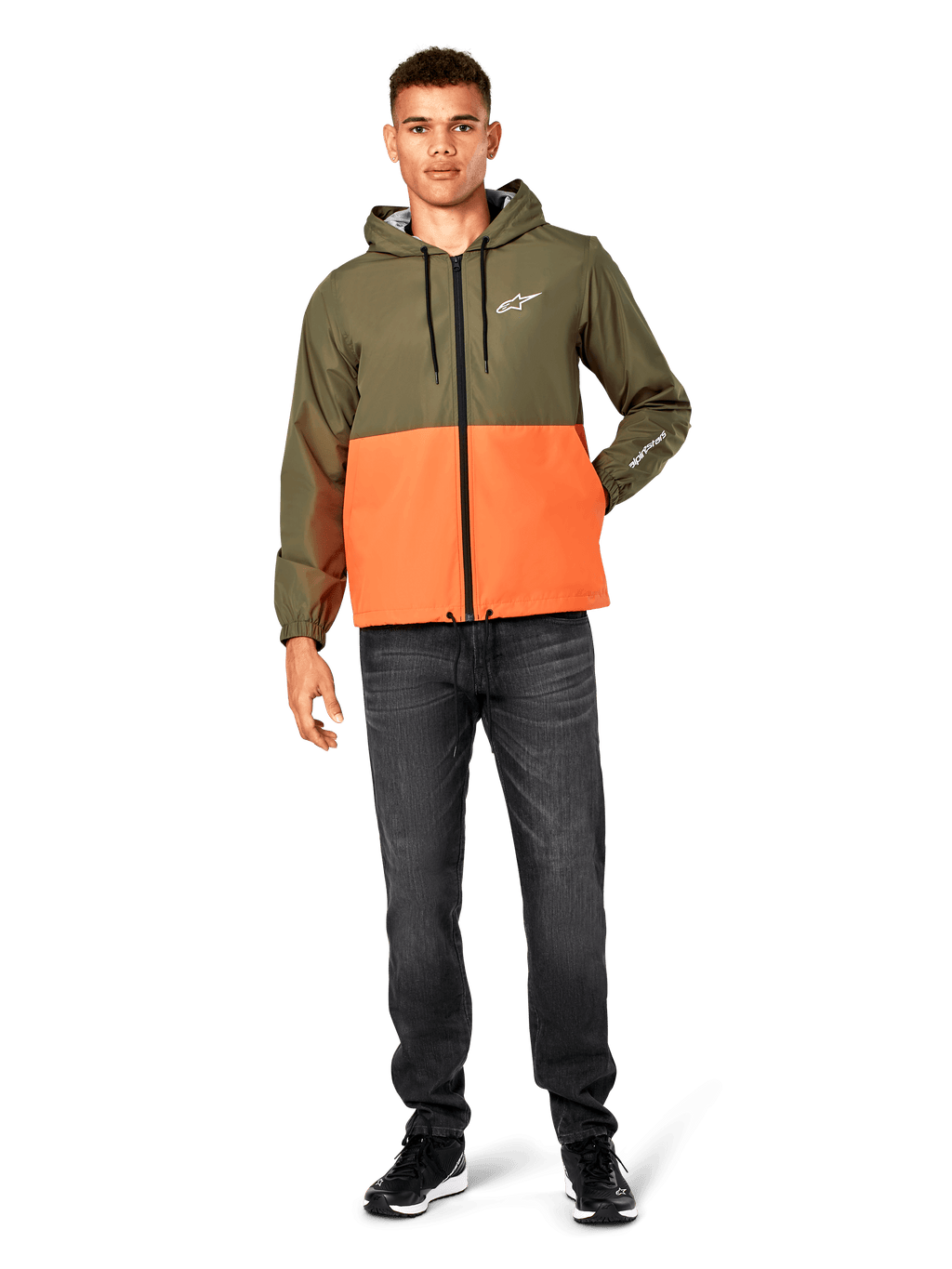 Chaqueta cortavientos Alpinestars Speeder, diseño en bloques de color verde militar y naranja, chaqueta ligera con capucha, cordones negros y cremallera frontal, logotipo blanco en el pecho y la manga, combinada con vaqueros oscuros y zapatillas negras, estilo deportivo casual.
