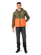 Chaqueta cortavientos Alpinestars Speeder, diseño en bloques de color verde militar y naranja, chaqueta ligera con capucha, cordones negros y cremallera frontal, logotipo blanco en el pecho y la manga, combinada con vaqueros oscuros y zapatillas negras, estilo deportivo casual.