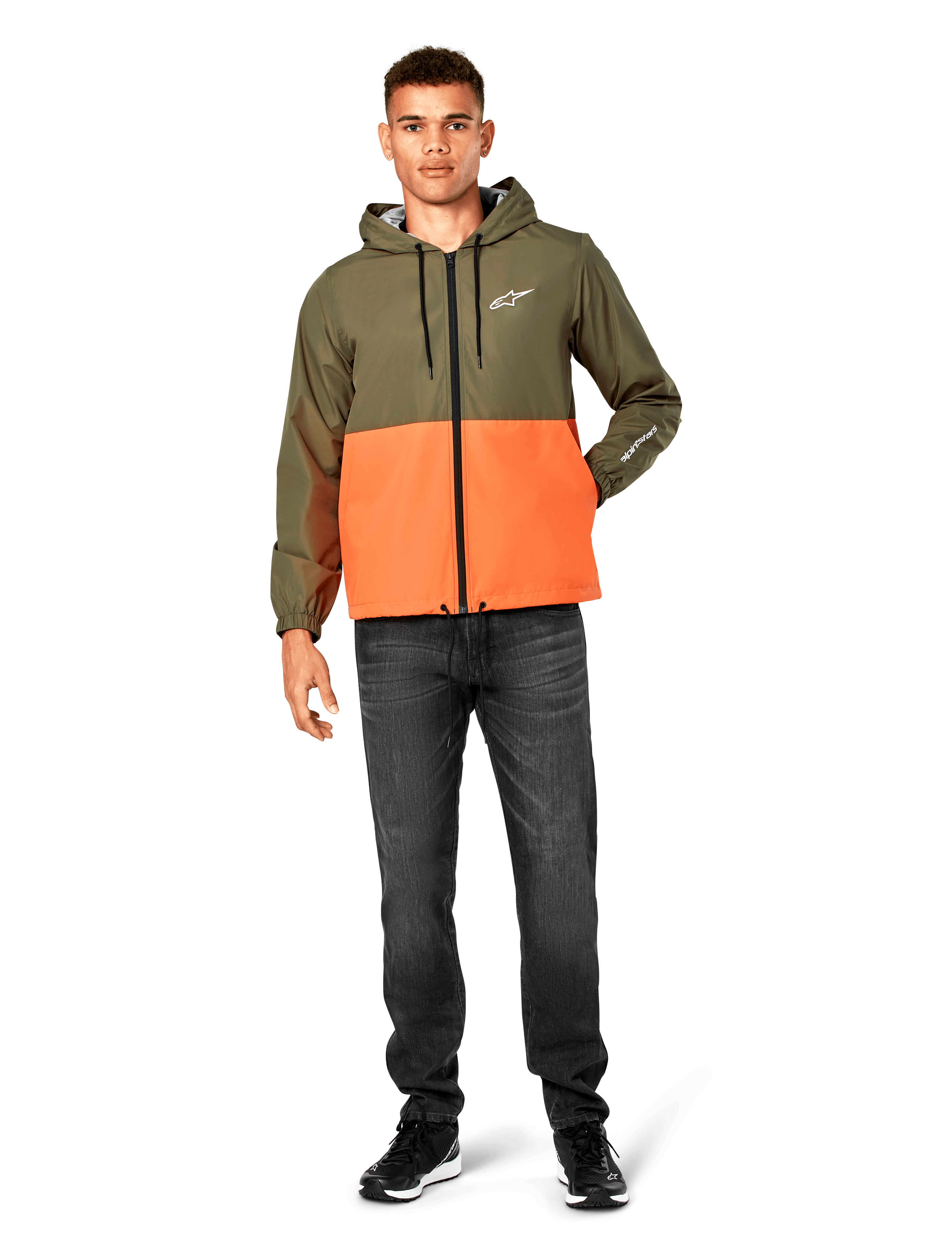 Chaqueta cortavientos Alpinestars Speeder, diseño en bloques de color verde militar y naranja, chaqueta ligera con capucha, cordones negros y cremallera frontal, logotipo blanco en el pecho y la manga, combinada con vaqueros oscuros y zapatillas negras, estilo deportivo casual.