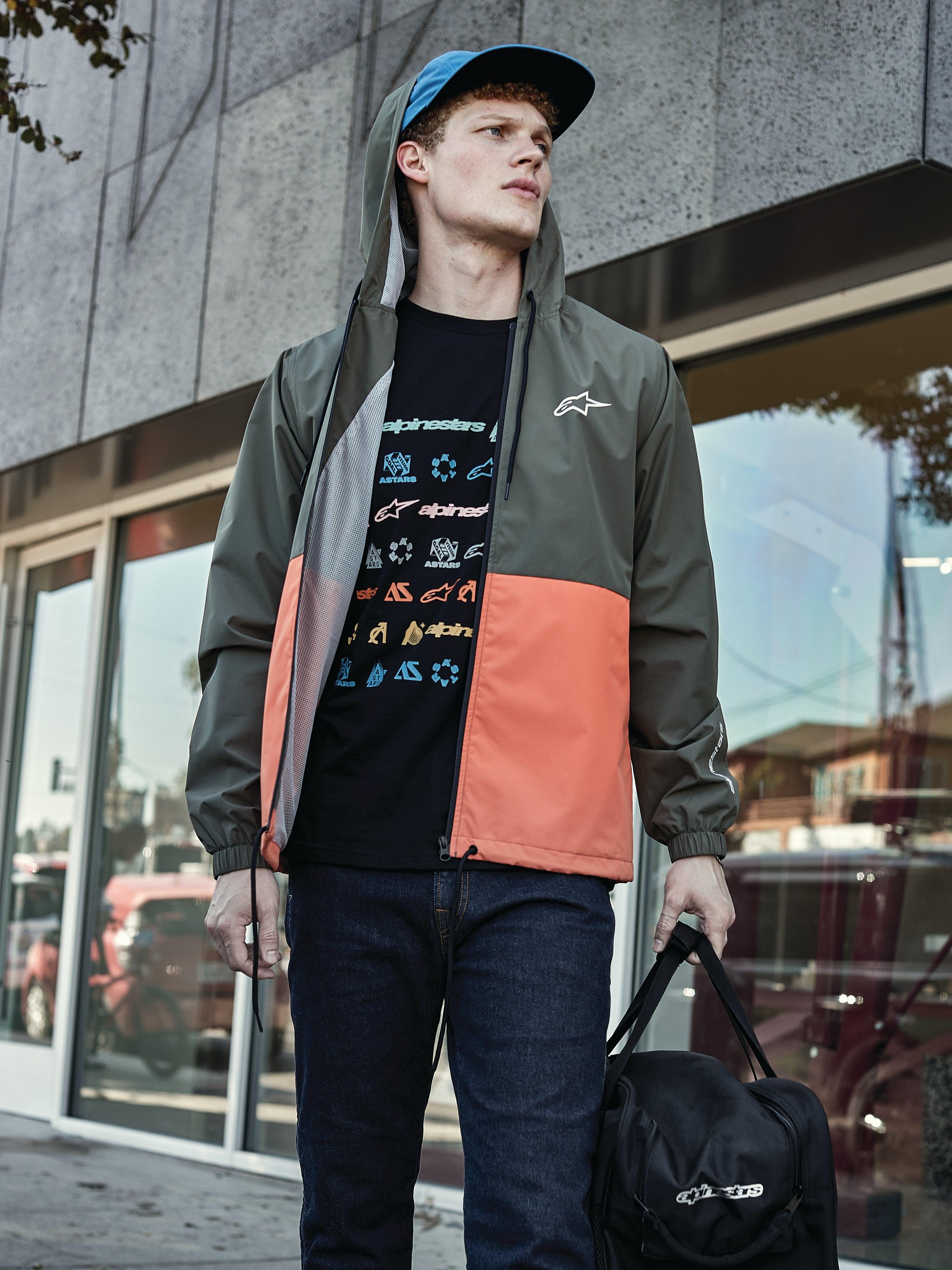 Hombre con la chaqueta cortavientos Alpinestars Speeder en Military Orange, con un diseño de bloques de color verde militar y naranja, capucha con forro de malla, combinada con una camiseta negra con logotipos gráficos, vaqueros azules y una bolsa de deporte Alpinestars negra, estilo de vida deportivo casual