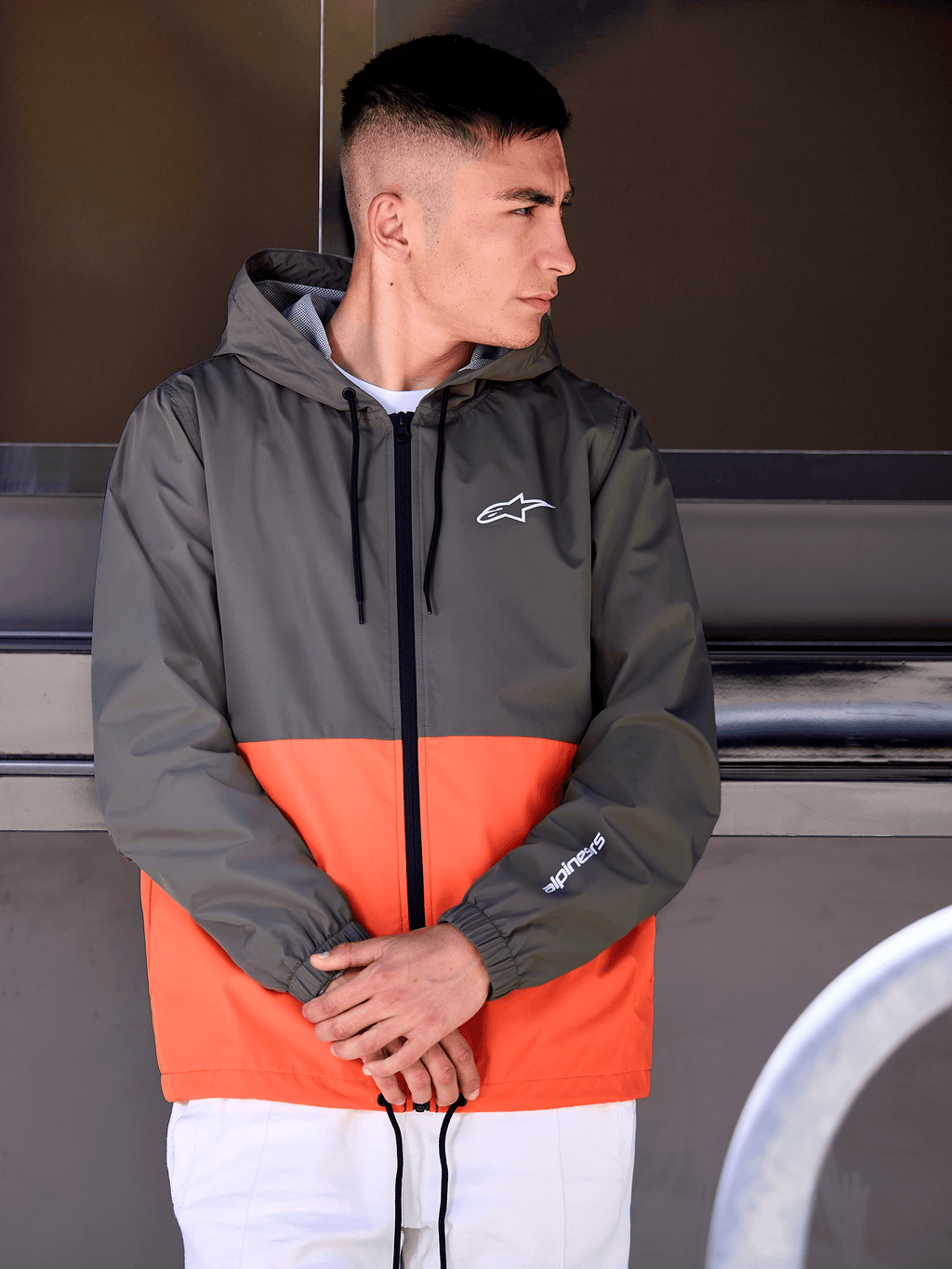 Hombre con la chaqueta cortavientos Speeder de Alpinestars, diseño de bloques de color en naranja militar y verde oscuro, prenda casual con capucha y logos blancos en el pecho y la manga, combinada con pantalones blancos para un look lifestyle inspirado en el motorsport