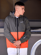 Hombre con la chaqueta cortavientos Speeder de Alpinestars, diseño de bloques de color en naranja militar y verde oscuro, prenda casual con capucha y logos blancos en el pecho y la manga, combinada con pantalones blancos para un look lifestyle inspirado en el motorsport