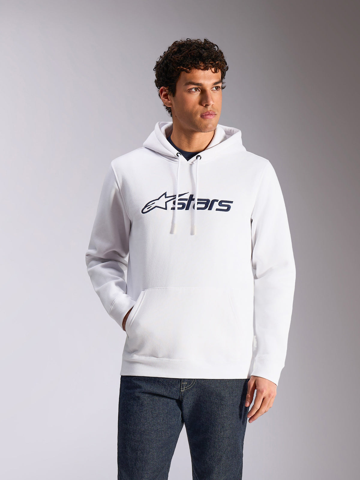 Sudadera Alpinestars Blaze V3, estilo urbano casual, blanca con logotipo Alpinestars azul marino, capucha con cordón, manga larga, combinada con pantalones vaqueros oscuros, diseño moderno inspirado en el automovilismo