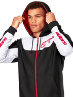 Alpinestars Sessions V3 Hoodie, negro, blanco y rojo, sudadera con cremallera frontal llevada por un hombre con la capucha puesta, con logotipos Alpinestars rojos en el pecho y las mangas, estilo streetwear casual de motorsport