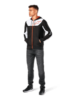Sudadera Alpinestars Sessions V3, sudadera casual con cremallera frontal, negro, blanco y rojo, lucida por un modelo con vaqueros oscuros y zapatillas negras, con paneles de bloques de color blanco en el pecho y las mangas, detalles de cremallera y cordón en rojo, logotipo grande en el pecho, ropa urbana de forro polar inspirada en el automovilismo