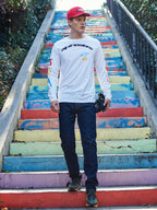 Alpinestars Brundo CSF Long Sleeve Tee, camiseta blanca de manga larga con logotipo negro de Alpinestars y coloridos estampados gráficos, lucida por un modelo en unas escaleras de colores, combinada con vaqueros azul oscuro y gorra roja, estilo streetwear casual de motorsport