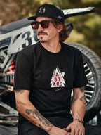 Alpinestars Gambit Trucker Hat in black with circular logo patch, worn with Alpinestars Soloist SS Tee in black, lifestyle shot featuring a man with sunglasses and tattoos sitting in front of a motocross bike.