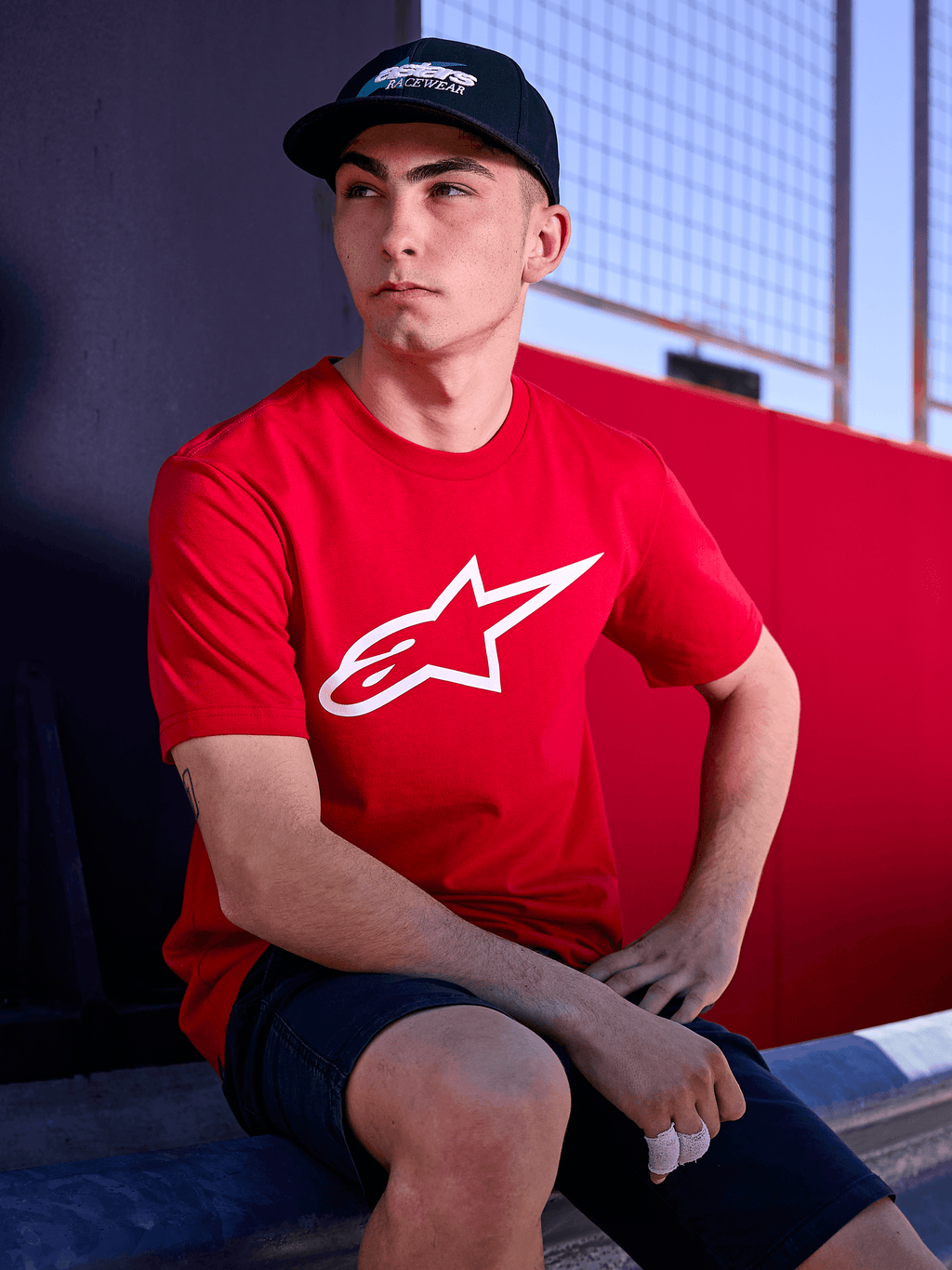 Gorra Alpinestars Profile en color negro, foto de estilo de vida de un hombre usando la gorra de visera curva con una camiseta roja con el logotipo de Alpinestars en blanco, estilo streetwear casual inspirado en el motor