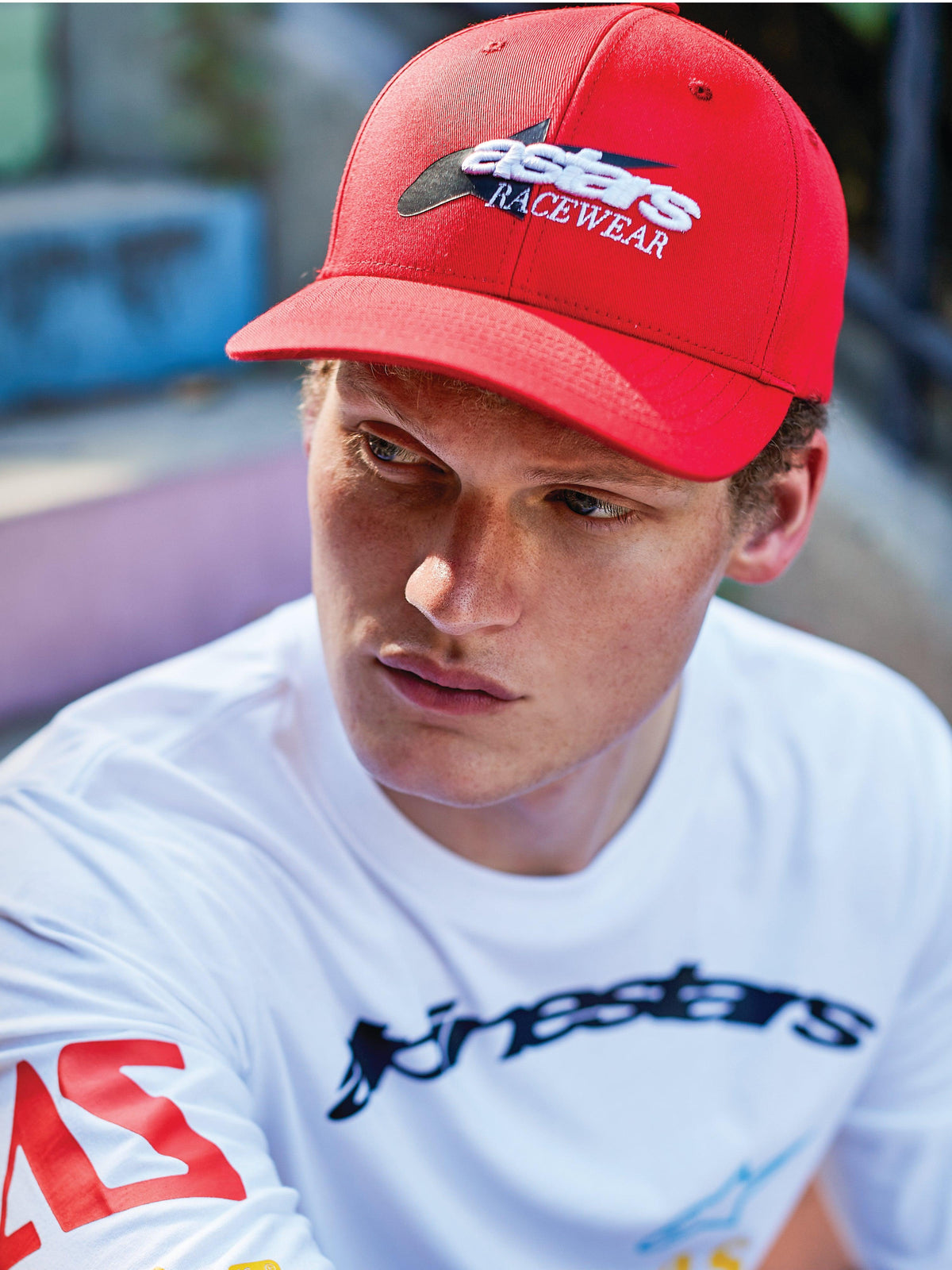 Hombre con gorra Profile de Alpinestars, snapback roja de visera curva con bordado frontal 'astars RACEWEAR' en blanco y negro, combinada con una camiseta blanca Alpinestars de manga larga con gráficos, estilo casual motorsport