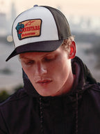 Gorra Trucker Alpinestars Hardware en Military White, lucida por un hombre con sudadera negra, con panel frontal blanco, visera curva y rejilla trasera negras, parche bordado en color arena con logotipo rojo de Alpinestars Motocross, moda urbana casual de automovilismo
