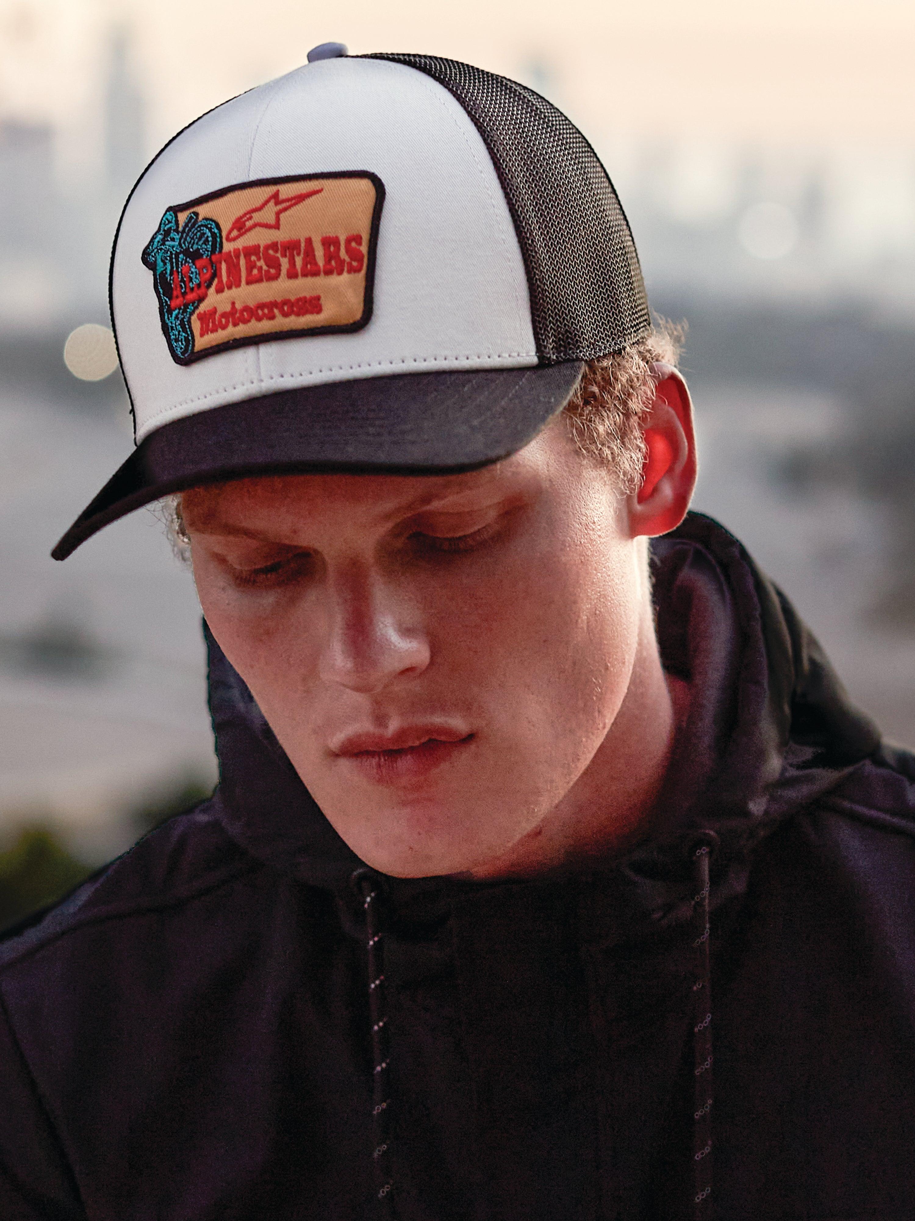 Gorra Trucker Alpinestars Hardware en Military White, lucida por un hombre con sudadera negra, con panel frontal blanco, visera curva y rejilla trasera negras, parche bordado en color arena con logotipo rojo de Alpinestars Motocross, moda urbana casual de automovilismo
