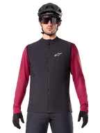 Alpinestars A-Dura Thermal Vest, chaleco de ciclismo térmico negro, usado por un modelo con un jersey granate de manga larga, casco de MTB negro y gafas de sol, con un logotipo blanco en el pecho, protección térmica ligera para ciclismo de montaña