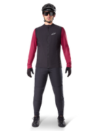 Alpinestars Chaleco Térmico A-Dura, chaleco de ciclismo negro sobre un jersey granate de manga larga, combinado con pantalones de MTB gris oscuro, casco y gafas de sol negras, ropa técnica de bicicleta de montaña para climas fríos
