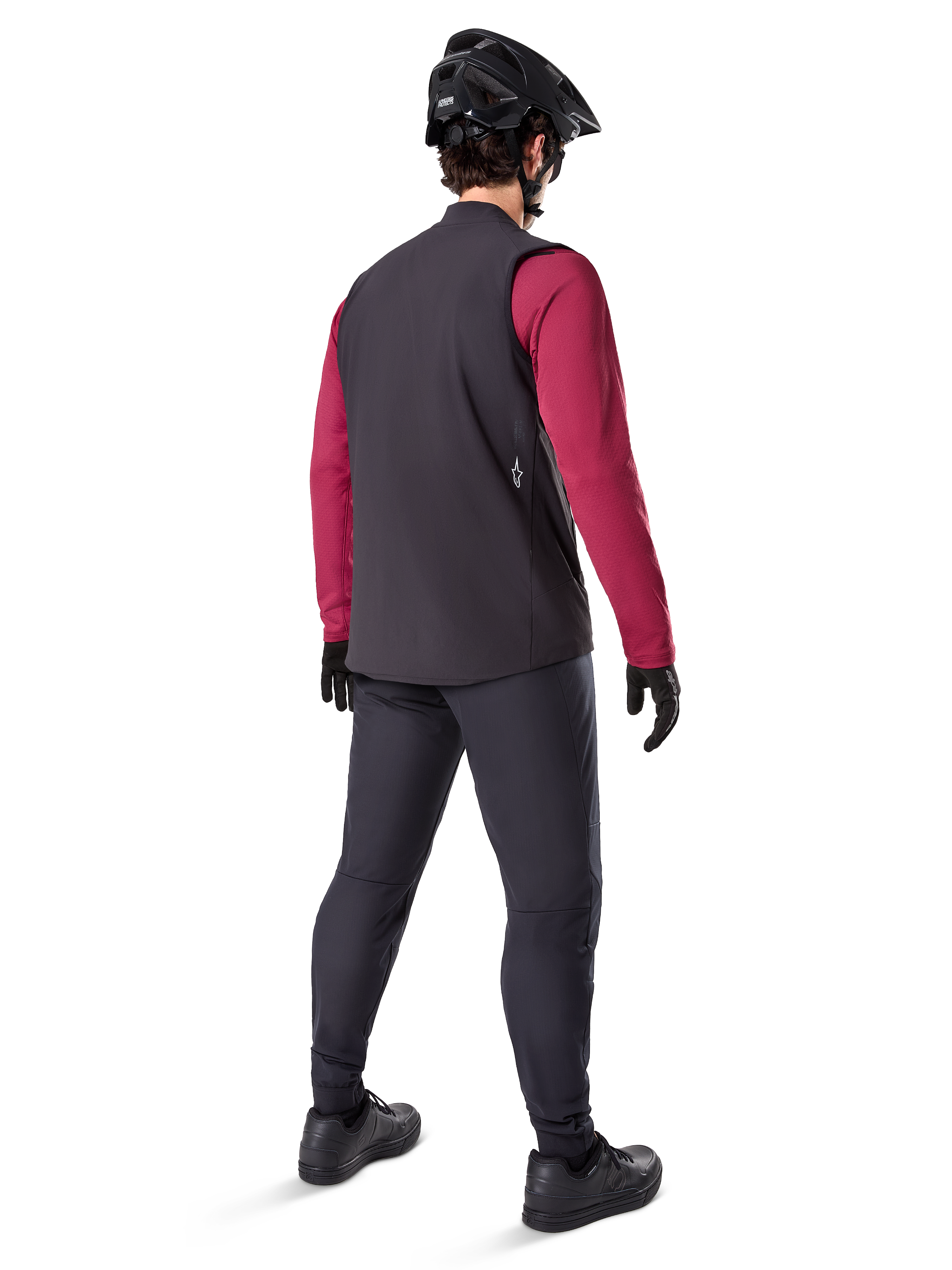 Chaleco Térmico Alpinestars A-Dura, chaleco de ciclismo negro, visto desde atrás en un modelo con un maillot granate de manga larga y pantalones de ciclismo negros, combinado con un casco de bicicleta de montaña y guantes negros, equipo técnico para rodar en climas fríos.