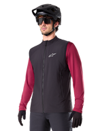 Alpinestars A-Dura Thermal Vest, chaleco térmico de ciclismo negro, usado sobre una camiseta de manga larga granate, con logotipo blanco en el pecho, combinado con casco de MTB negro y gafas de sol, capa de rendimiento con aislamiento para ciclismo de montaña