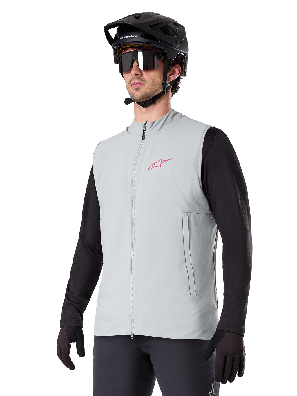 Alpinestars A-Dura Thermal Vest, gris Titanium, chaleco térmico de ciclismo, usado sobre una camiseta negra de manga larga con casco de bicicleta de montaña y guantes negros, con un logo rosa en el pecho y bolsillos laterales con cremallera vertical