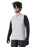 Alpinestars A-Dura Thermal Vest, gris Titanium, chaleco térmico de ciclismo, usado sobre una camiseta negra de manga larga con casco de bicicleta de montaña y guantes negros, con un logo rosa en el pecho y bolsillos laterales con cremallera vertical