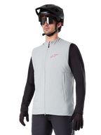 Alpinestars A-Dura Thermal Vest, gris Titanium, chaleco térmico de ciclismo, usado sobre una camiseta negra de manga larga con casco de bicicleta de montaña y guantes negros, con un logo rosa en el pecho y bolsillos laterales con cremallera vertical