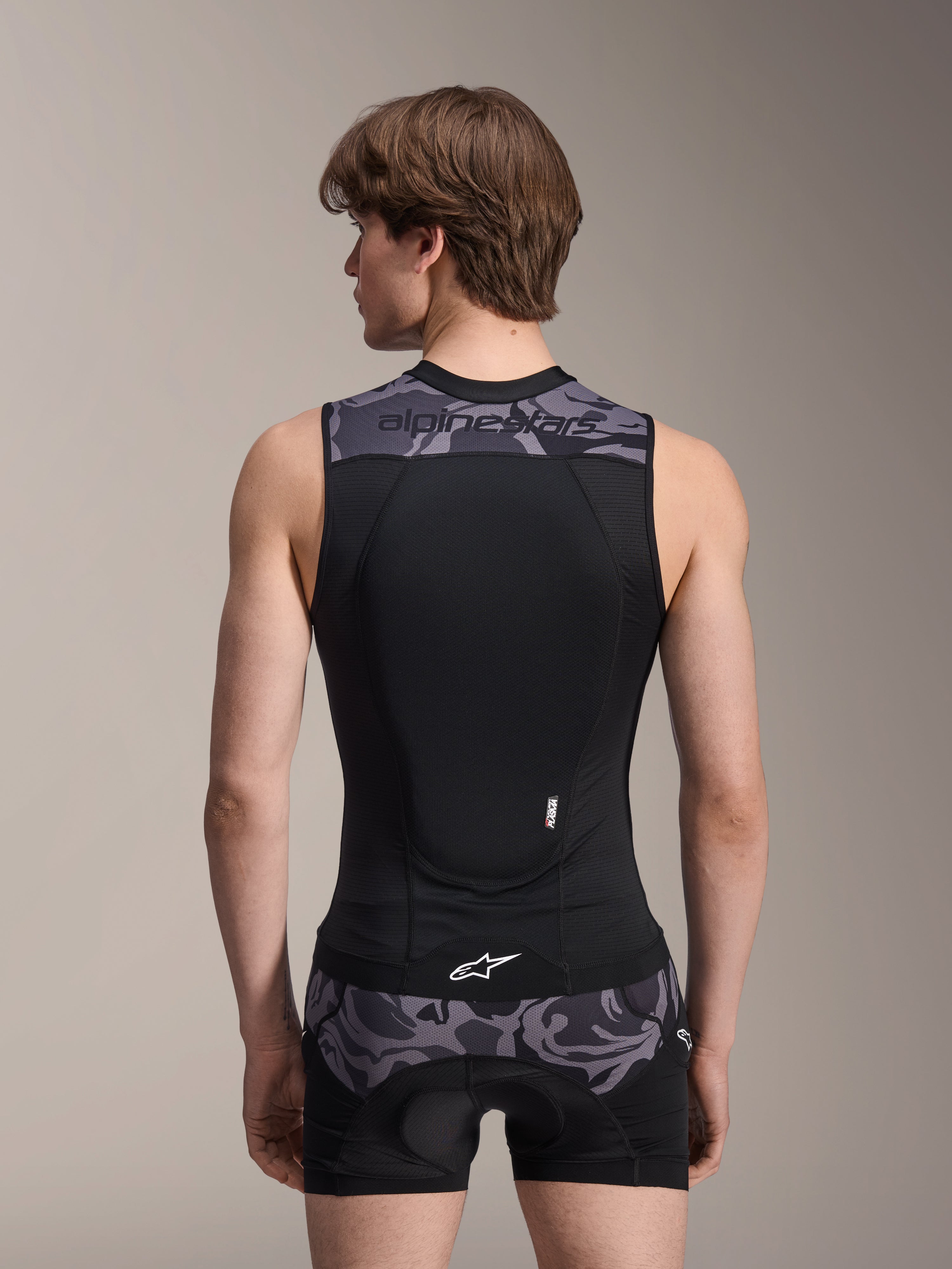 Chaleco de protección Alpinestars A-Motion Plasma, Black Camo, vista trasera en modelo masculino, con construcción de malla transpirable, protector de espalda Nucleon Plasma integrado, detalles de camuflaje gris en los hombros y la cintura, y logotipo de Alpinestars.