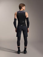 Chaleco de protección Alpinestars A-Motion Plasma, vista trasera, negro con detalles de camuflaje gris en los hombros, usado por un modelo con coderas negras, pantalones cortos de ciclismo sobre mallas y zapatillas negras, destacando el protector de espalda integrado
