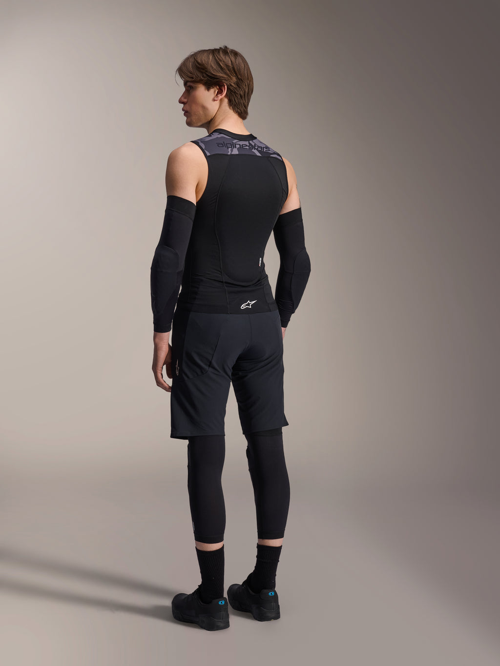 Modelo masculino de espaldas vistiendo el Chaleco de Protección Alpinestars A-Motion Plasma, Black Camo, diseño de malla sin mangas con protector de espalda integrado, patrón de camuflaje gris en los hombros, usado con coderas negras y pantalones cortos de ciclismo de montaña, equipamiento de ciclismo profesional