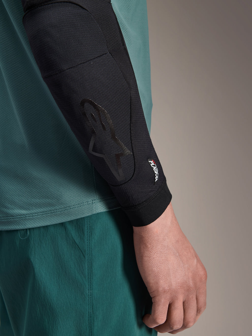 Alpinestars A-Motion Plasma Elbow Sleeve, codera de MTB negra, vista de primer plano puesta en el brazo con jersey verde azulado, con el logo de Astars tonal y la marca Nucleon Plasma, diseño de malla de perfil delgado para ciclismo todoterreno
