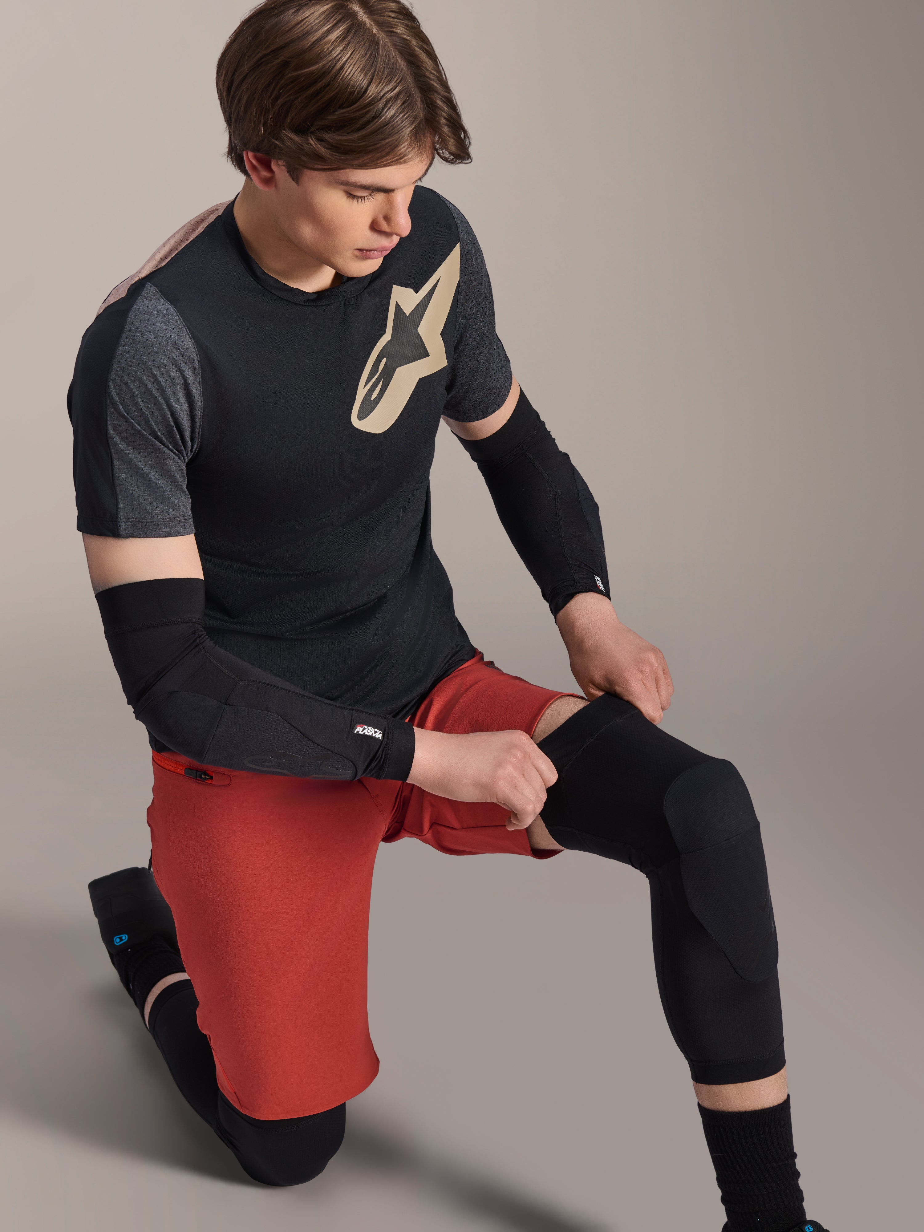Alpinestars A-Motion Plasma Knee Sleeve, rodillera de MTB, negro, usada por un ciclista arrodillado, combinada con una camiseta negra y gris con logotipo dorado, pantalones cortos rojos y coderas negras, equipo de protección profesional para bicicleta de montaña