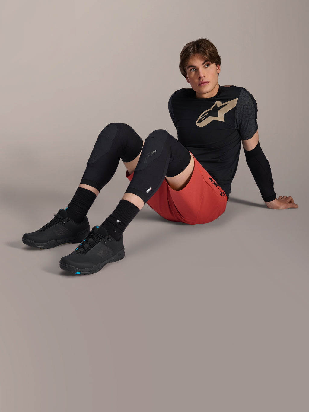 Codera Alpinestars A-Motion Plasma, usada por un ciclista masculino sentado en el suelo de un estudio, con construcción de malla negra, combinada con pantalones cortos rojos y un jersey Alpinestars negro con logotipo beige, protección de alto rendimiento para ciclismo de montaña y trail.