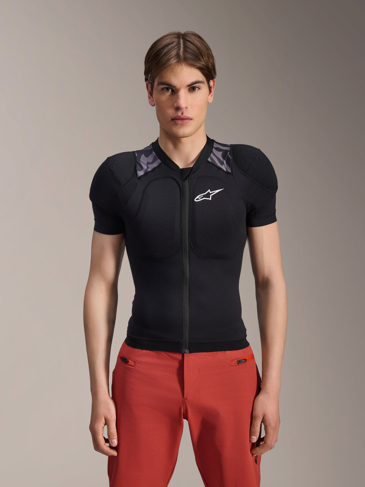 Modelo masculino vistiendo la chaqueta de protección de manga corta Alpinestars A-Motion Plasma, color negro con acolchado de camuflaje gris en los hombros, logotipo de Alpinestars blanco en el pecho, combinada con pantalones rojos, armadura corporal de MTB de ajuste ceñido con cremallera frontal