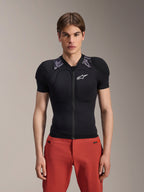Modelo masculino vistiendo la chaqueta de protección de manga corta Alpinestars A-Motion Plasma, color negro con acolchado de camuflaje gris en los hombros, logotipo de Alpinestars blanco en el pecho, combinada con pantalones rojos, armadura corporal de MTB de ajuste ceñido con cremallera frontal